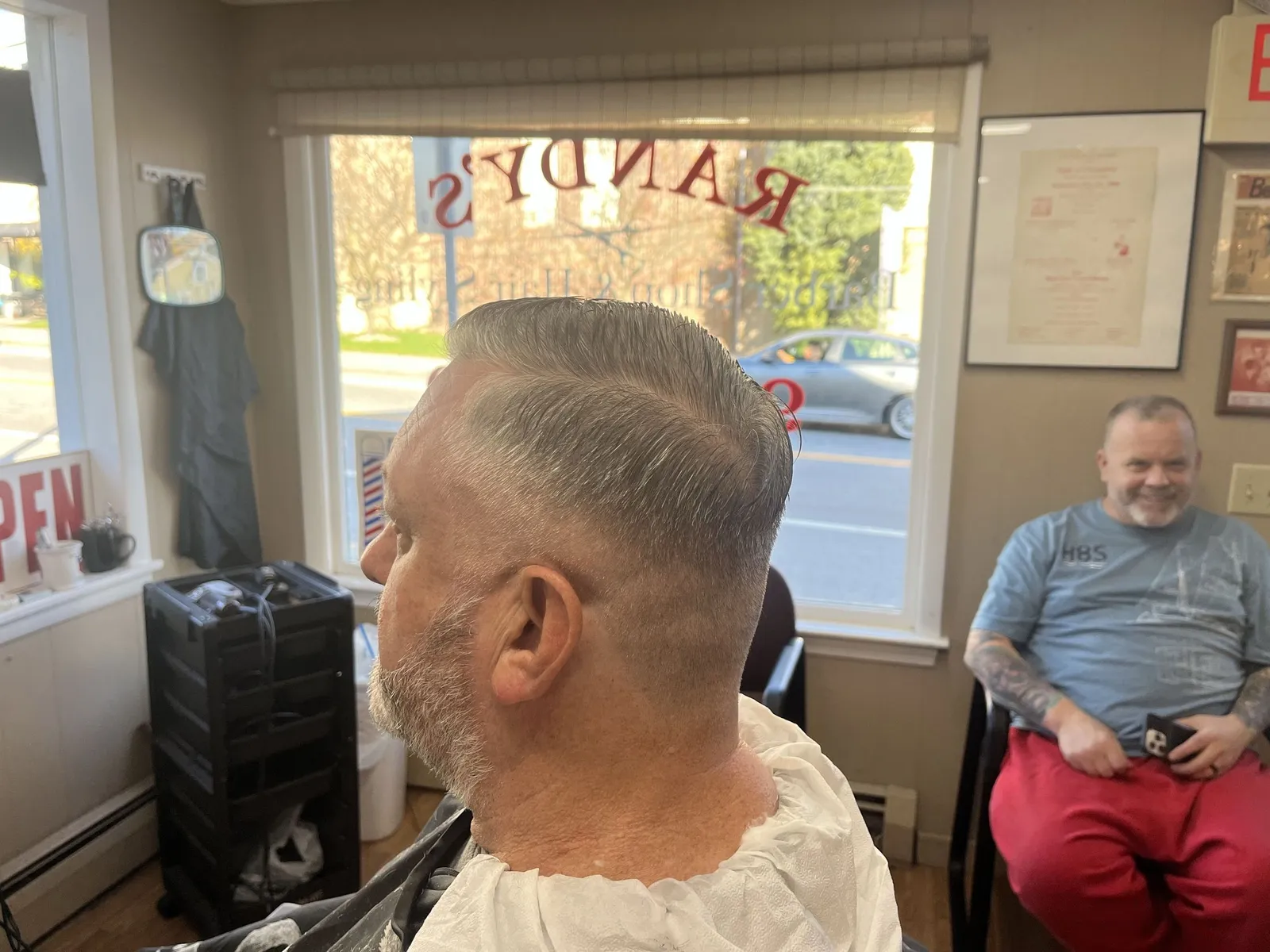 Side profile of a grey-haired man with his hair slicked back and a low taper, the RANDY'S window lettering behind him