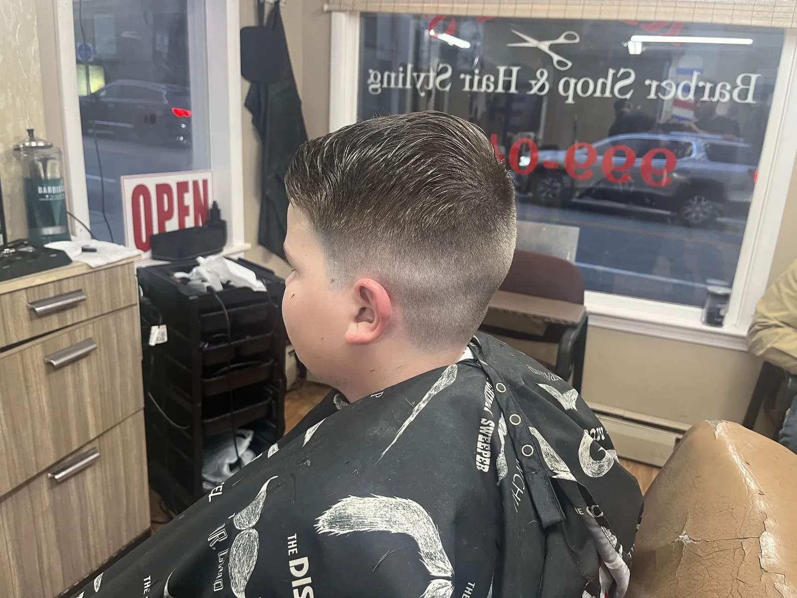 A boy with a combed-over top and a mid skin fade, in front of the Barber Shop & Hair Styling window