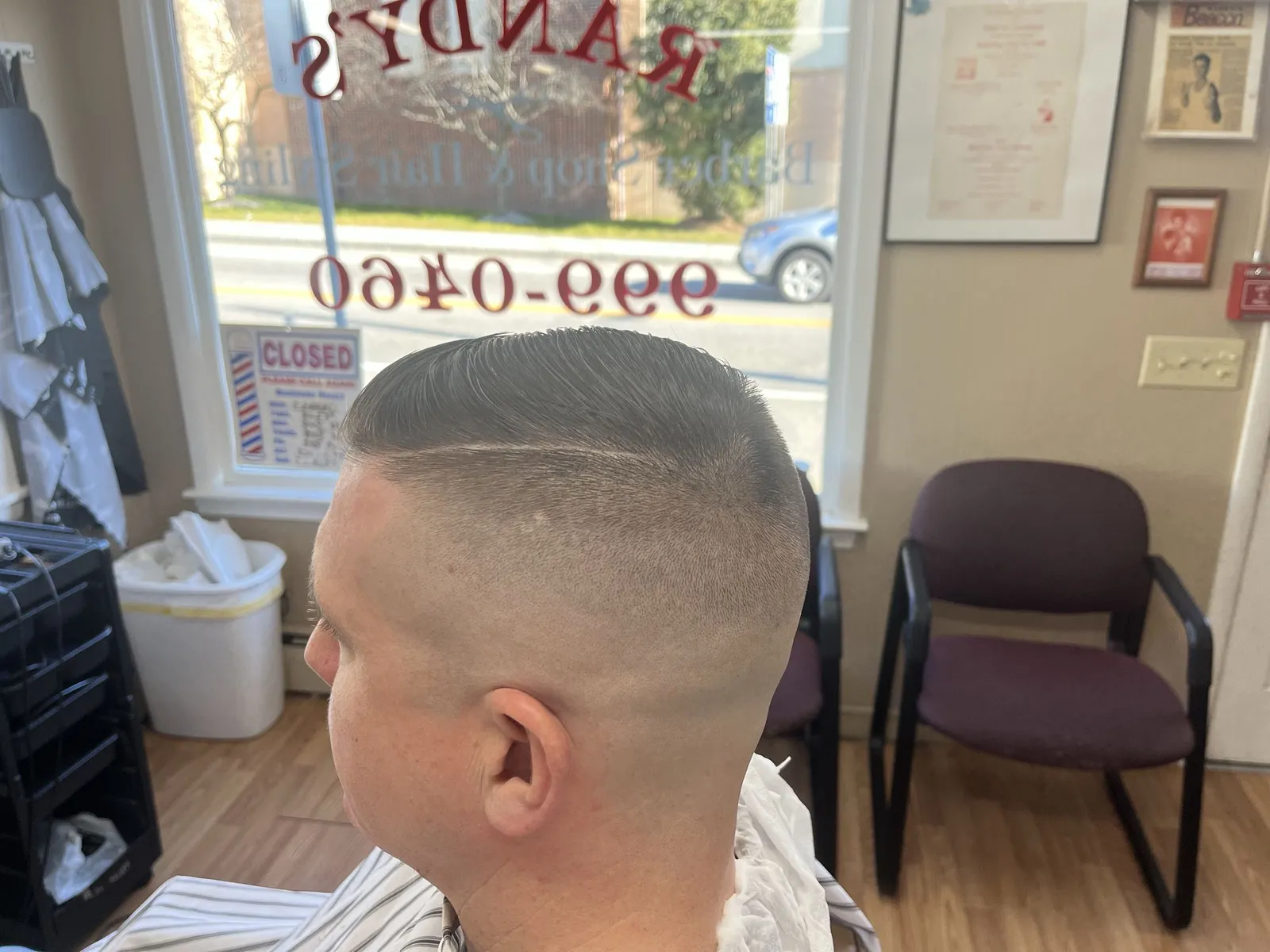 Side profile of a man with a hard part and a high skin fade, the shop's phone number on the window behind